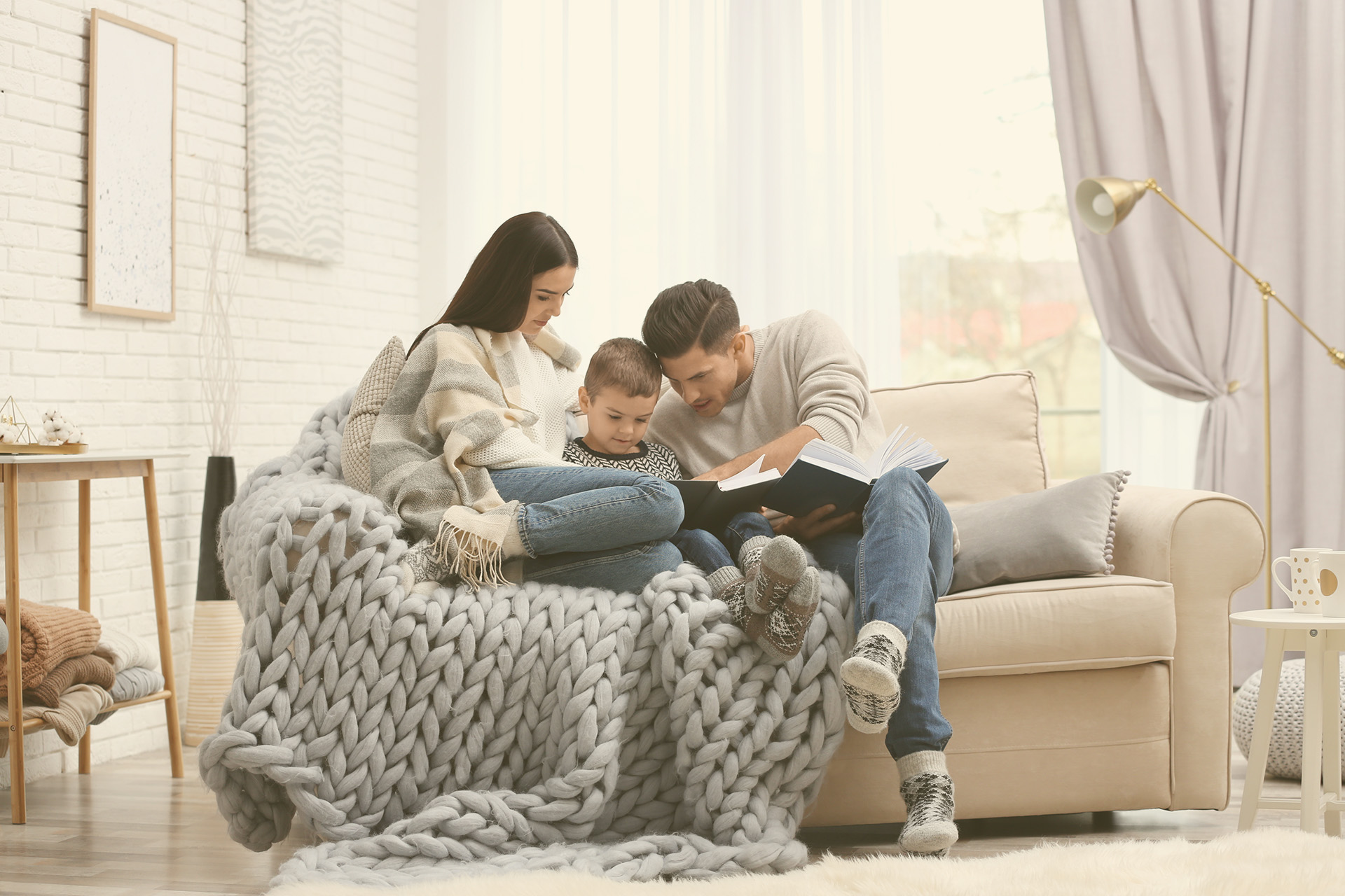A young family reads on a small couch. This image is for an article about how to prepare for a winter storm in an apartment. The article includes tips on gathering emergency supplies, how to keep perishable foods safe and more.